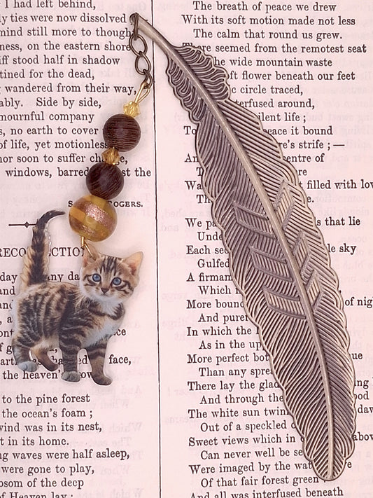 Decorative flat feather bookmark with  topaz color bead and wooden beads and tabby kitten charm a textured paper background 