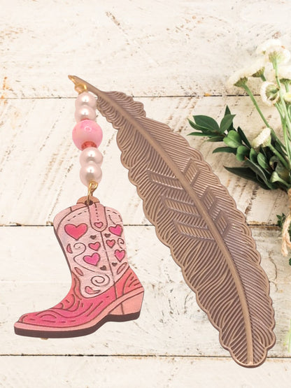 Pink cowboy boot ornament with heart patterns, pearl beads, and feather brass-tone bookmark on a wooden surface with flowers.