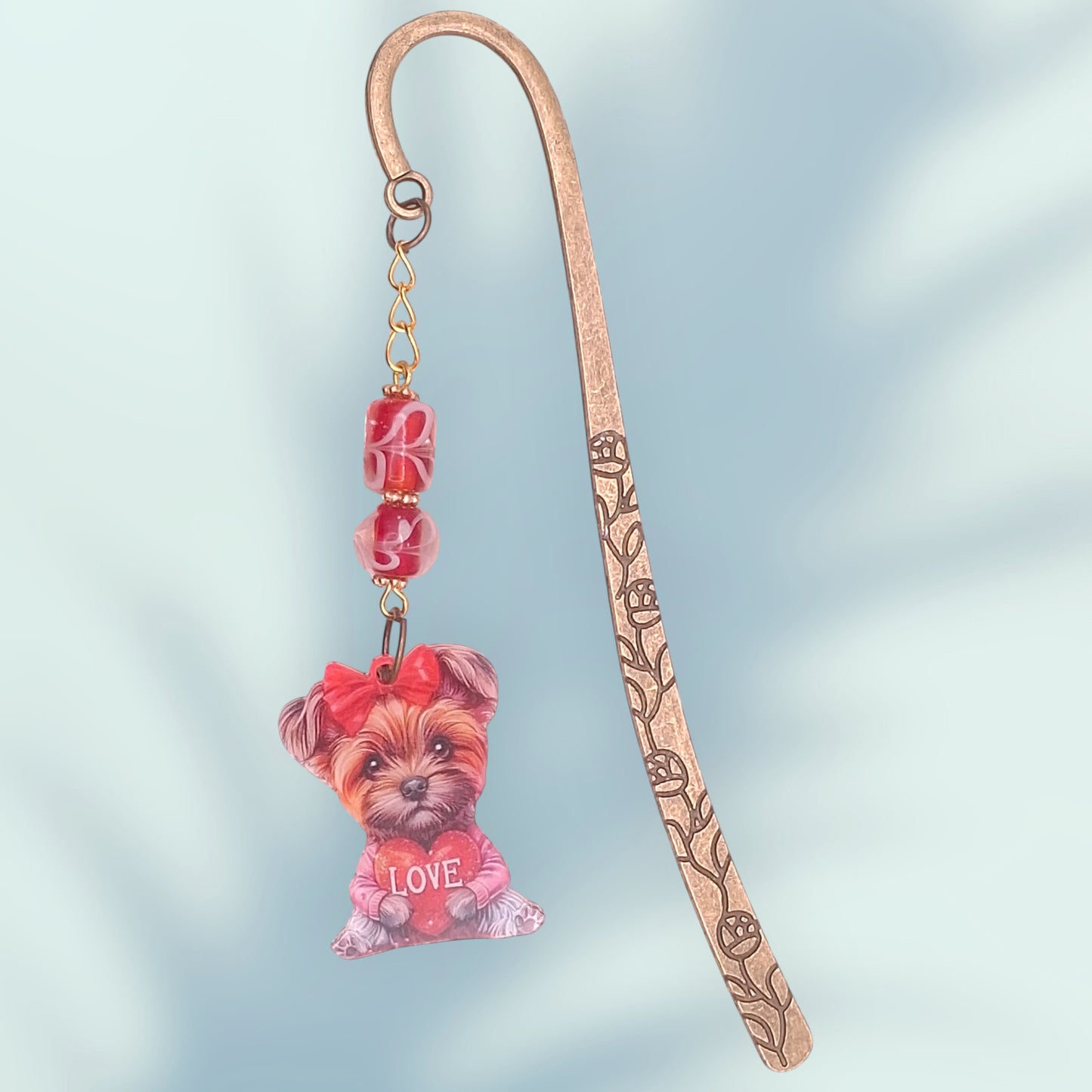 Brass-tone floral etched bookmark with a puppy terrier charm with the work LOVE and red decorative beads on a light blue background
