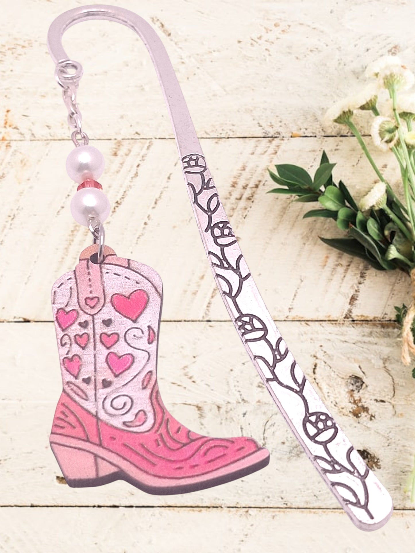 Pink cowboy boot on silver-tone bookmark with heart designs on a white wooden surface with flowers.