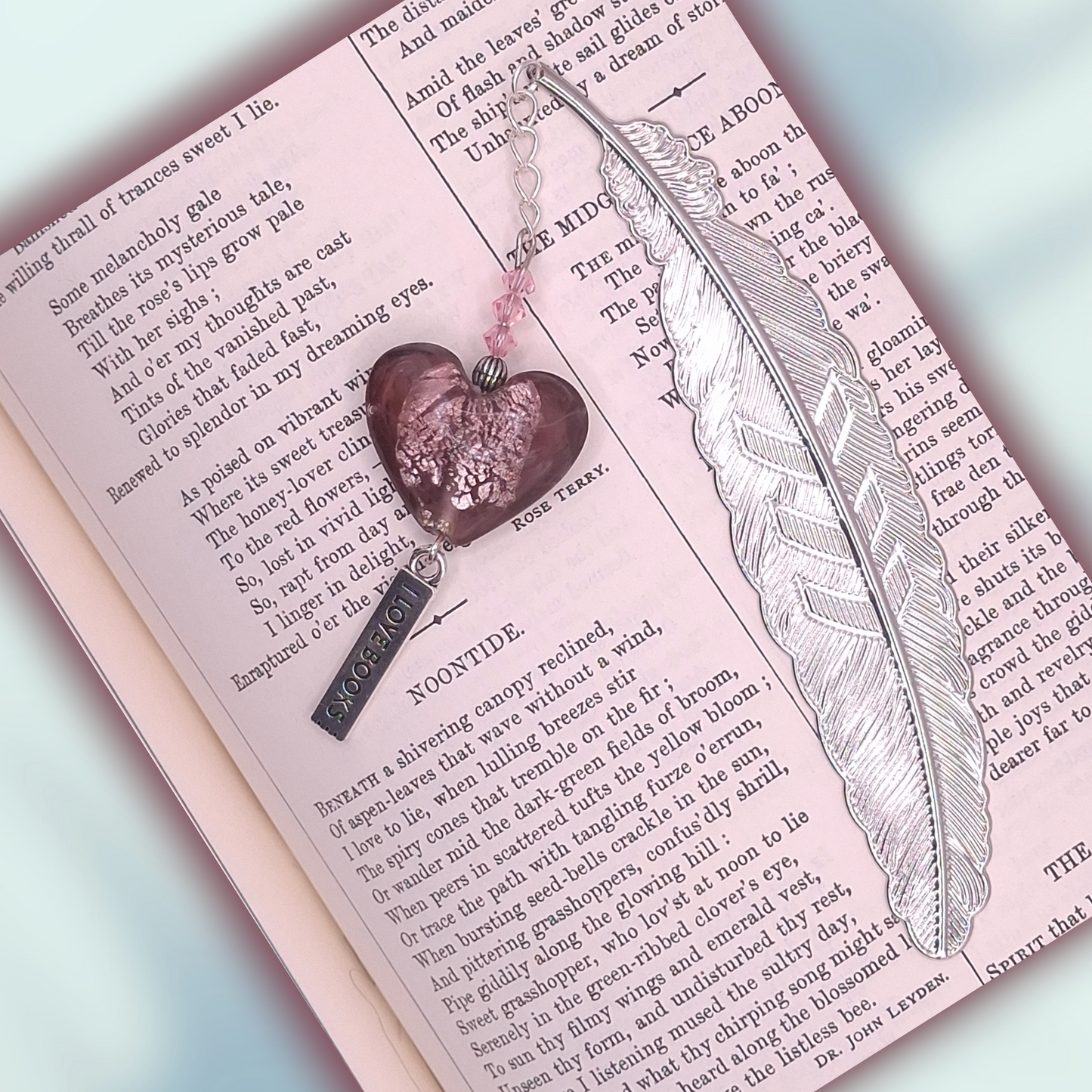 Silver-tone flat feather bookmark with accented with pink bicones and plum heart-shaped charm with rose iridescent crackling in the center, and with 'I Love Books' charm on a decorative background with text.