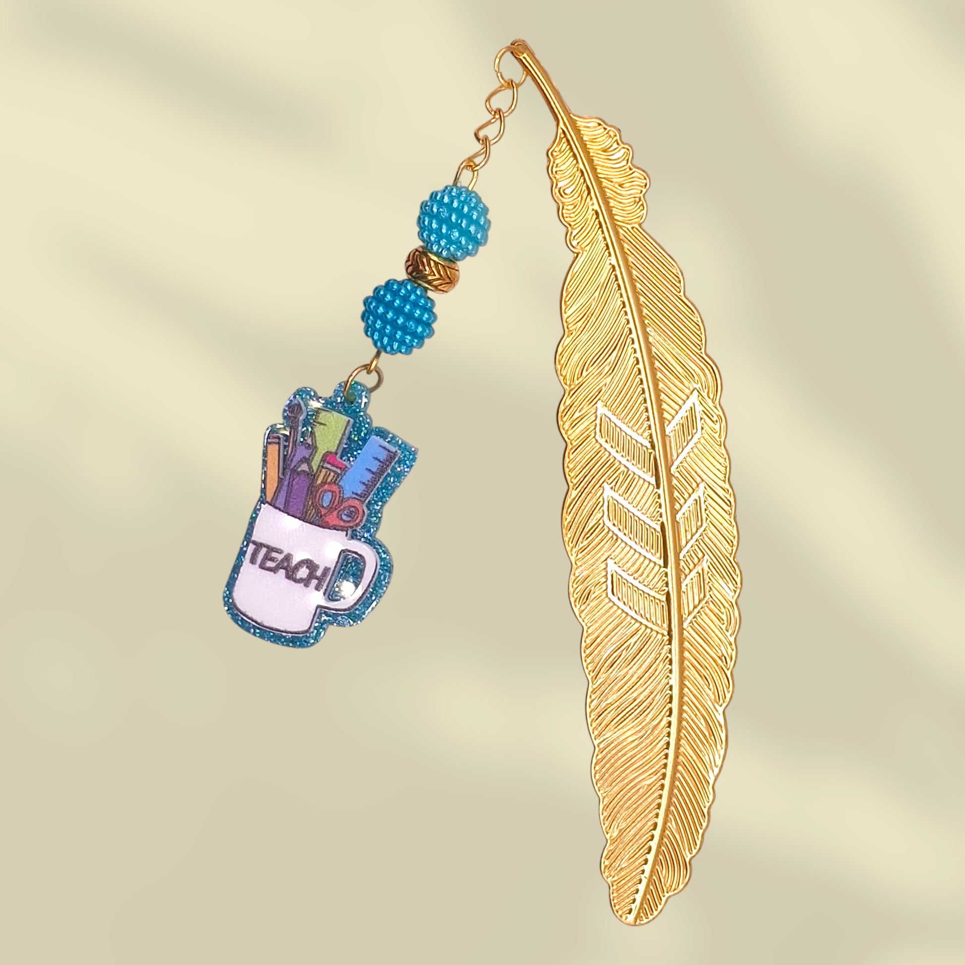 Gold feather-shaped bookmark with blue bayberry beads and cup with the word teach. Inside the cup are school tools - ruler, scissor, pen, pencil, etc.