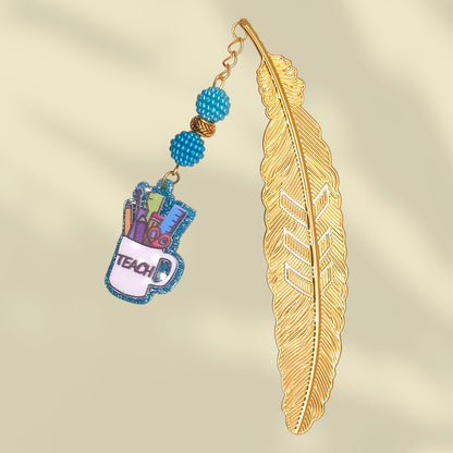 Gold feather-shaped bookmark with blue bayberry beads and cup with the word teach. Inside the cup are school tools - ruler, scissor, pen, pencil, etc.
